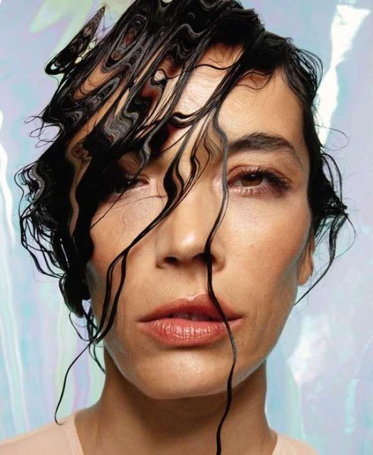 Close-up portrait of an adult woman with wet black hair against an iridescent background, showcasing beauty and fashion.