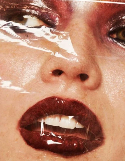 Close-up portrait of a female face covered in plastic wrap, featuring red lipstick and detailed eye makeup. Beauty and fashion aesthetic.