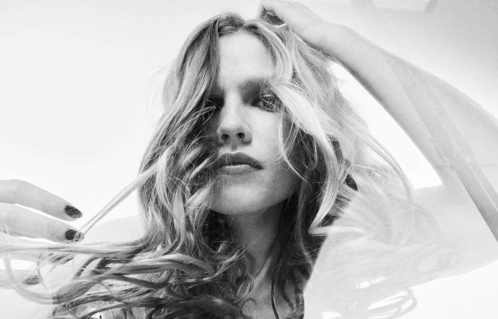 Monochrome close-up of a blonde woman with dark nails, touching her hair against a bright white backdrop, creating a stark and modern portrait.