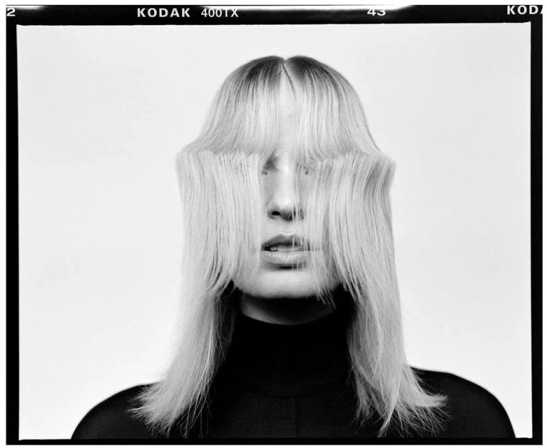 Fashion shot of a woman with blonde hair covering her face, wearing a black turtleneck sweater, against a white background in a photo studio.