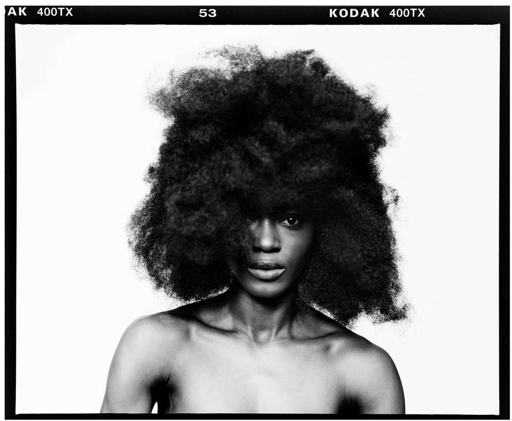 Striking monochrome portrait of a Black woman with an afro, showcasing her face and upper torso against a stark white background with film border.