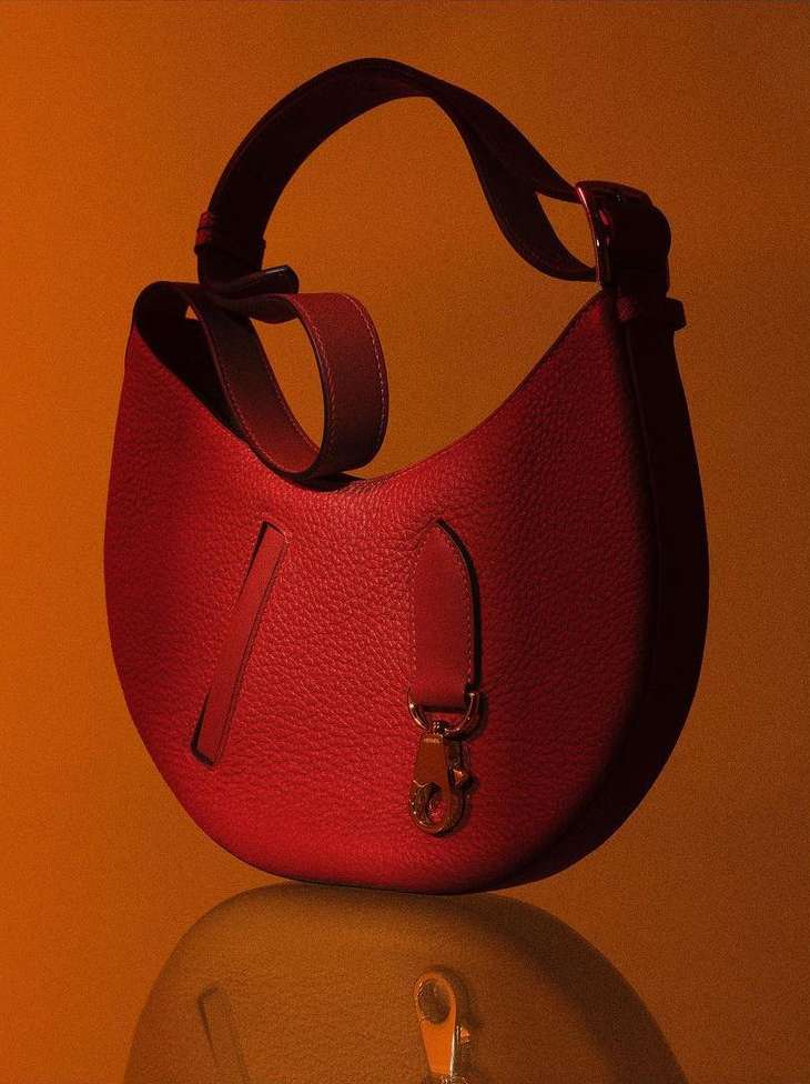 Studio shot of a red leather purse with a shoulder strap and metal hardware, sitting on a reflective surface with a warm orange backdrop.