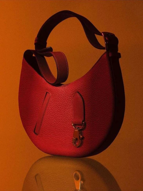 Studio shot of a red leather purse with a shoulder strap and metal hardware, sitting on a reflective surface with a warm orange backdrop.