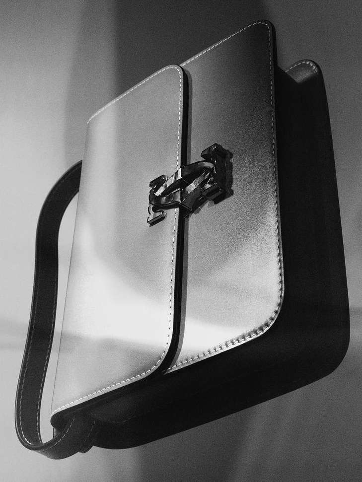 Black and white photograph of a modern handbag with a silver clasp. High-contrast image focuses on the bag's structured shape and textures.