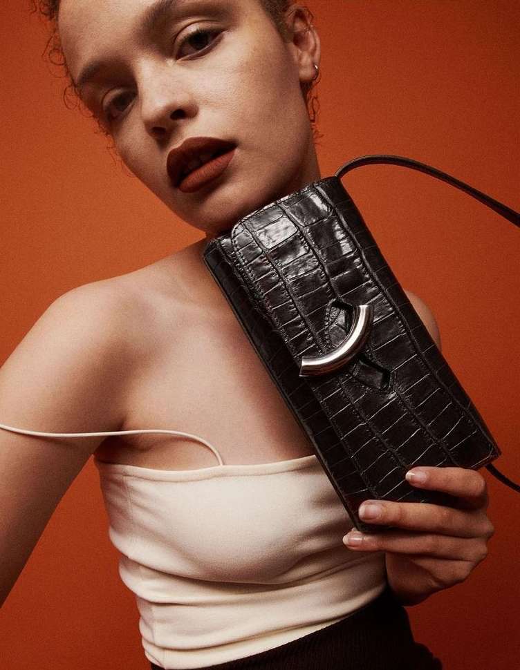 Portrait of a woman holding a black crocodile-skin purse against an orange background, showcasing fashion and beauty.