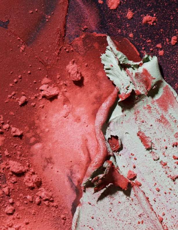 Close-up of coral pink and pale green cosmetic powder, with a gritty texture, create an abstract pattern against a dark background.