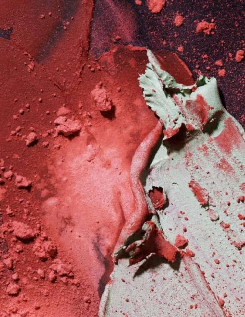 Close-up of coral pink and pale green cosmetic powder, with a gritty texture, create an abstract pattern against a dark background.