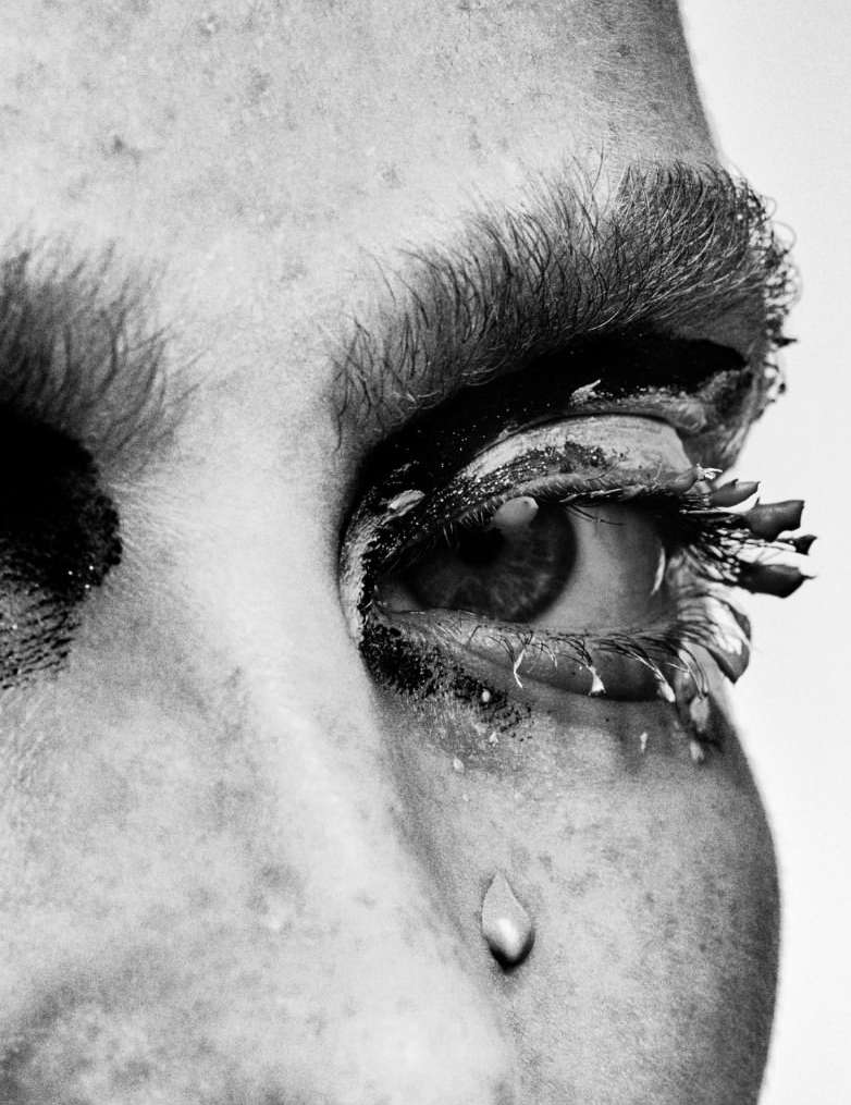 Close-up black and white portrait of a person's face with a tear dropping from their eye and heavy makeup on their eyelashes and eyebrows.
