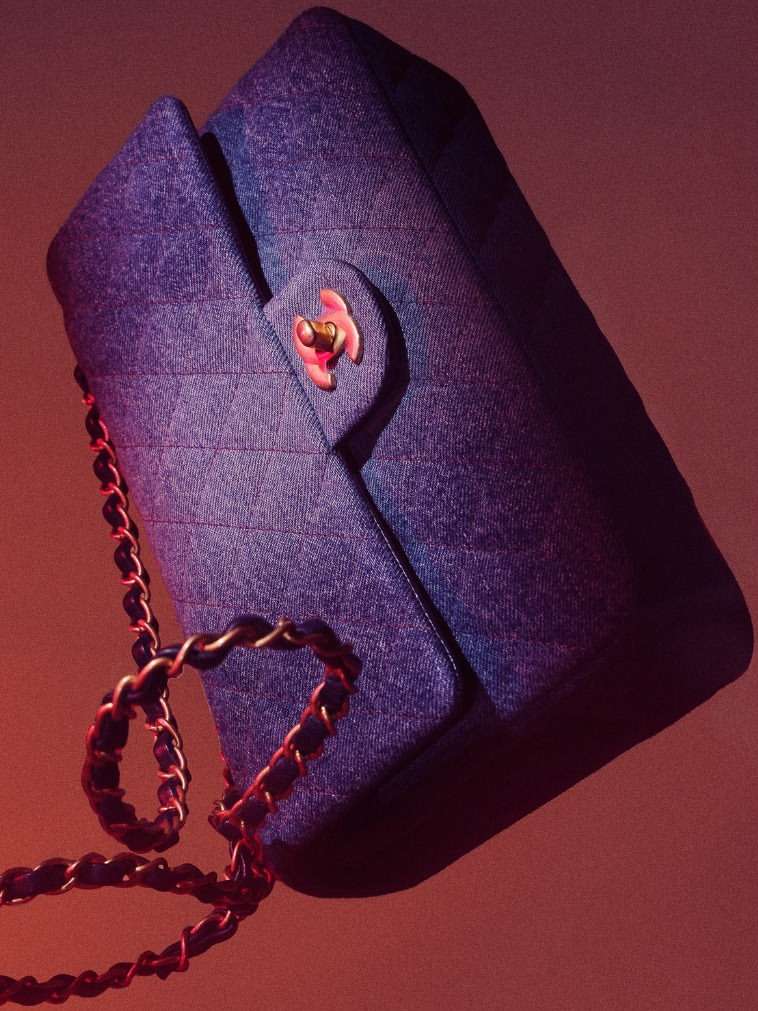 Close-up studio shot of a denim Chanel flap bag with a gold chain strap and pink clasp against a gradient pink-peach background.