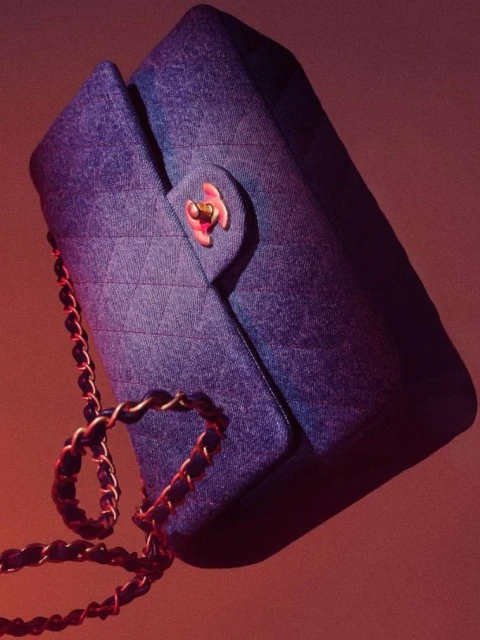 Close-up studio shot of a denim Chanel flap bag with a gold chain strap and pink clasp against a gradient pink-peach background.