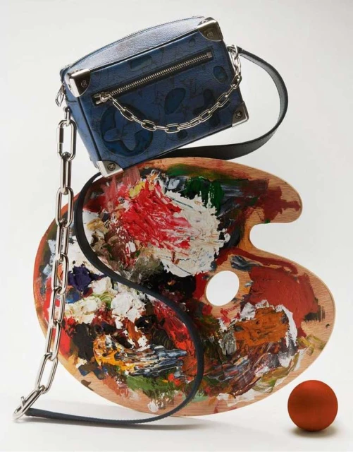 Luxury fashion still life with blue Louis Vuitton purse balancing on a wooden paint palette next to a red sphere on white.