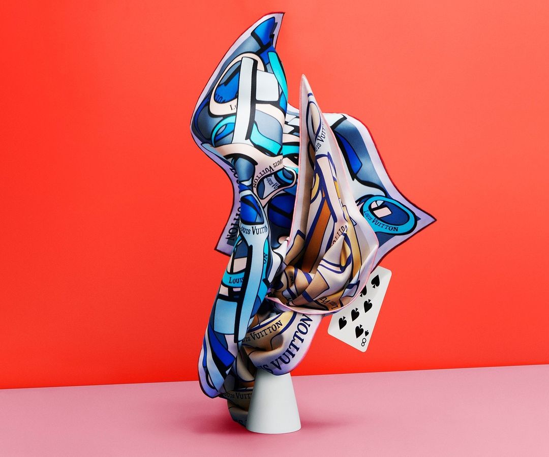 Fashion still life featuring a patterned silk scarf arranged on a white pedestal, accented by a playing card against a red backdrop.