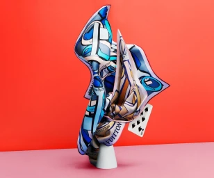 Scrunched blue and white silk scarf draped on a white stand with a playing card against a coral backdrop.