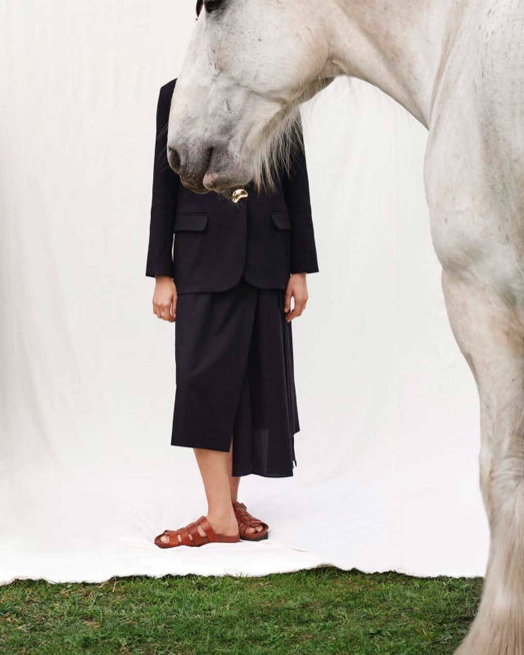Fashion editorial shot of a woman wearing a black suit and sandals, standing alongside the head of a white horse, set outdoors.
