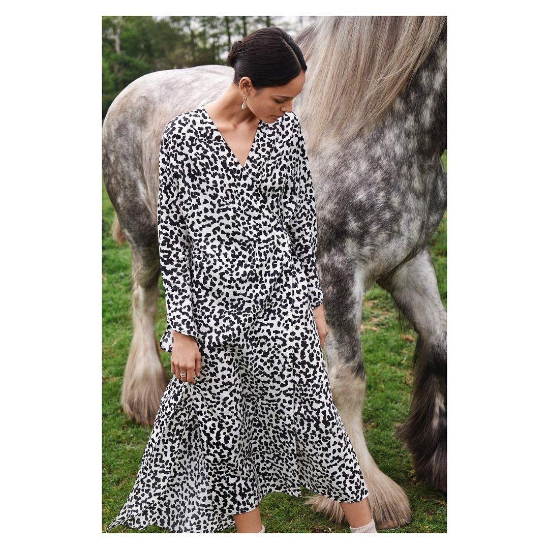 Adult woman in a printed long-sleeve maxi dress posing next to a grey horse in a grassy outdoor setting with trees.