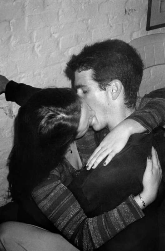 Black and white photo of a couple kissing indoors, with a brick wall visible in the background and the woman wearing a striped shirt.
