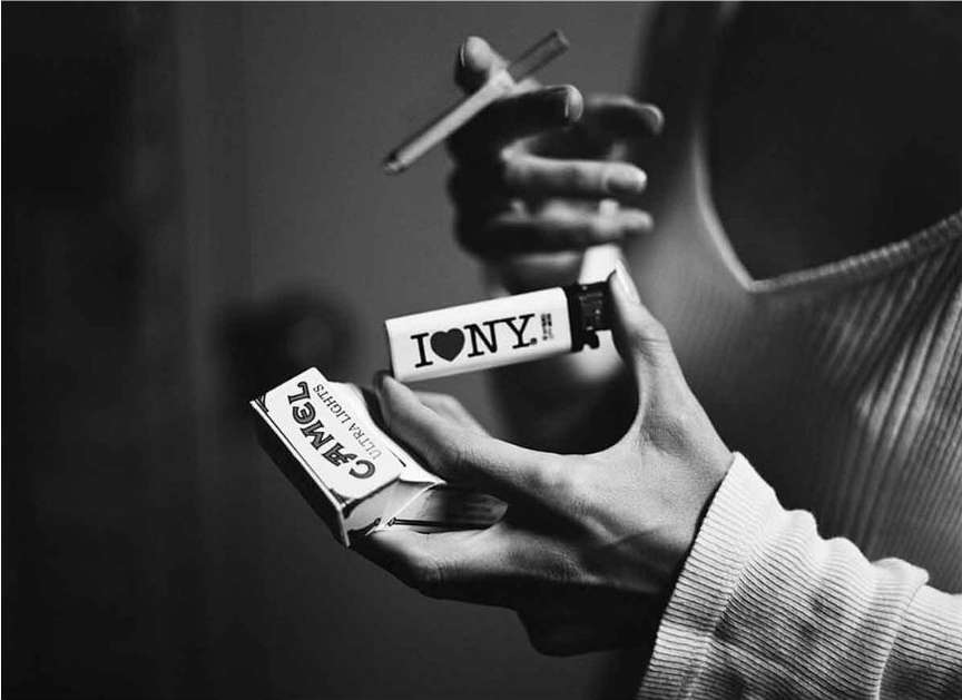 Close-up shot of hands holding a cigarette, lighter with an 'I LOVE NY' graphic, and a pack of Camel Ultra Lights in black and white.