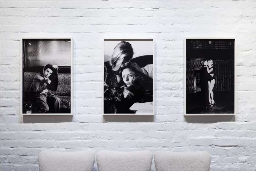 Three black and white framed photographic prints featuring people are displayed on a white brick wall above modern, light grey chairs.