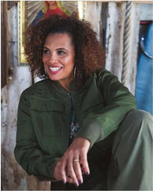 Smiling woman with curly hair wears a green jacket and sits indoors with a painting in the background, creating a warm, inviting portrait.