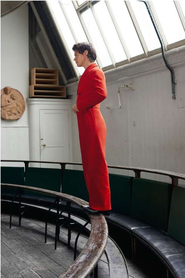 Adult male wearing a red suit stands on a railing in a brightly lit, vintage-style indoor space with tiered seating and architectural details.