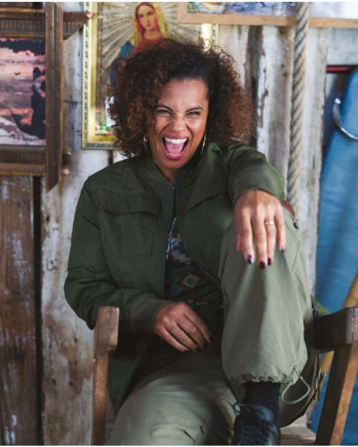 Excited woman with curly hair in a green jacket and trousers sitting on a wooden chair in a rustic indoor setting with pictures on the wall.