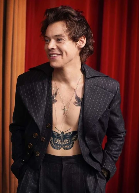 A portrait of smiling man in a pinstripe suit, showcasing tattoo art on his chest, against a warm-toned studio backdrop.