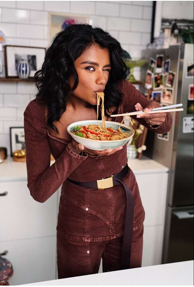 Attractive woman in brown outfit eating noodles with chopsticks. She's indoors in a kitchen with a partial view of the interior.
