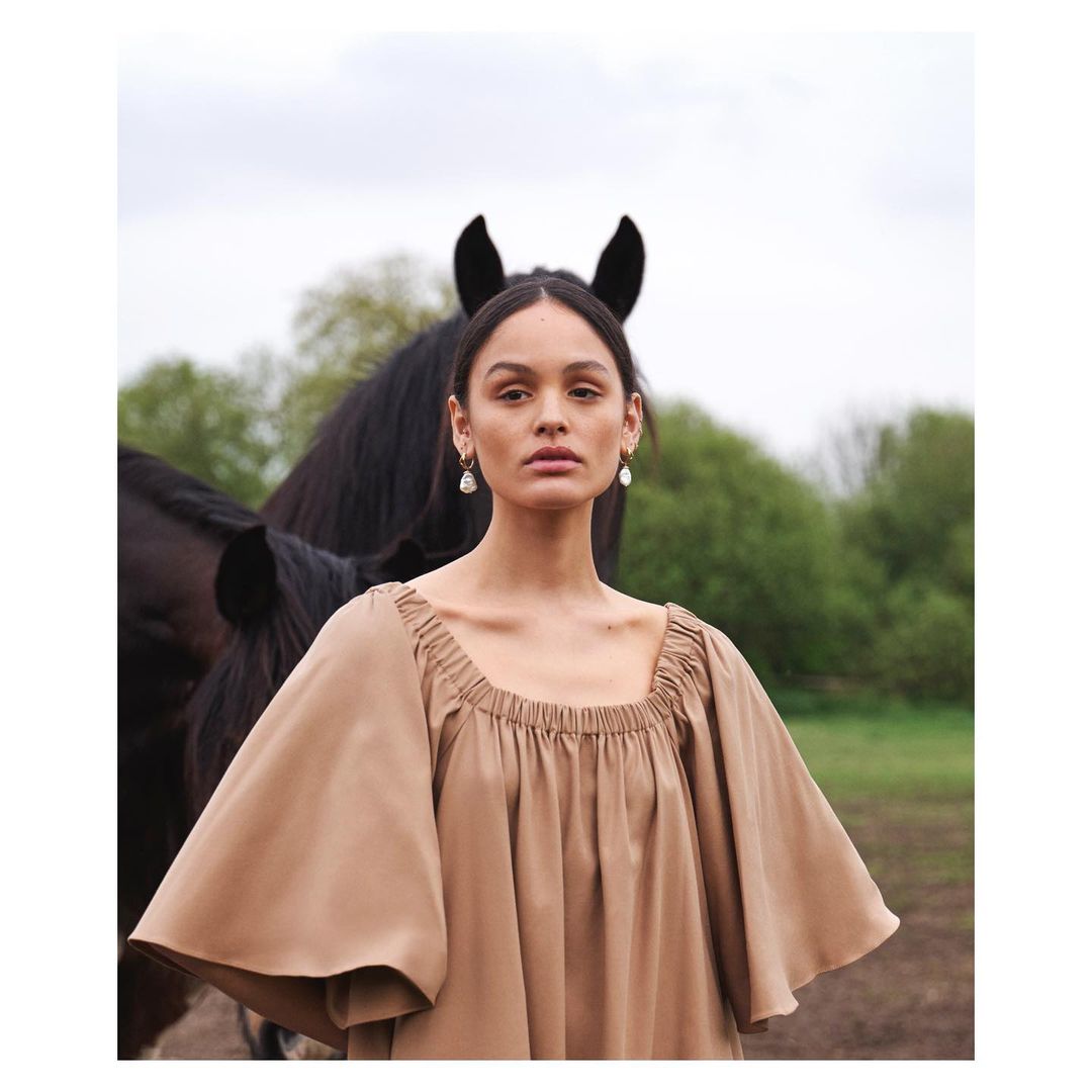 Portrait of a woman in a tan dress, standing outdoors next to a dark horse with trees in the background, soft natural lighting.