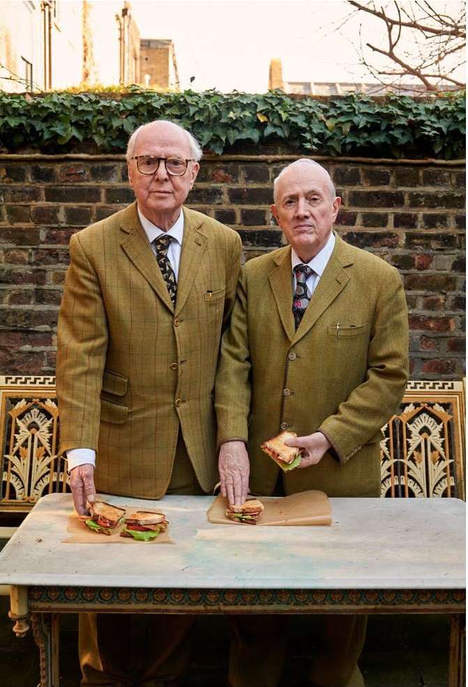 Two elderly men in matching tweed jackets stand outside behind sandwiches. They wear glasses and ties, and green ivy climbs a brick wall.