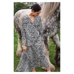 Woman in black and white patterned dress standing beside a dapple-grey horse outdoors in a grassy field