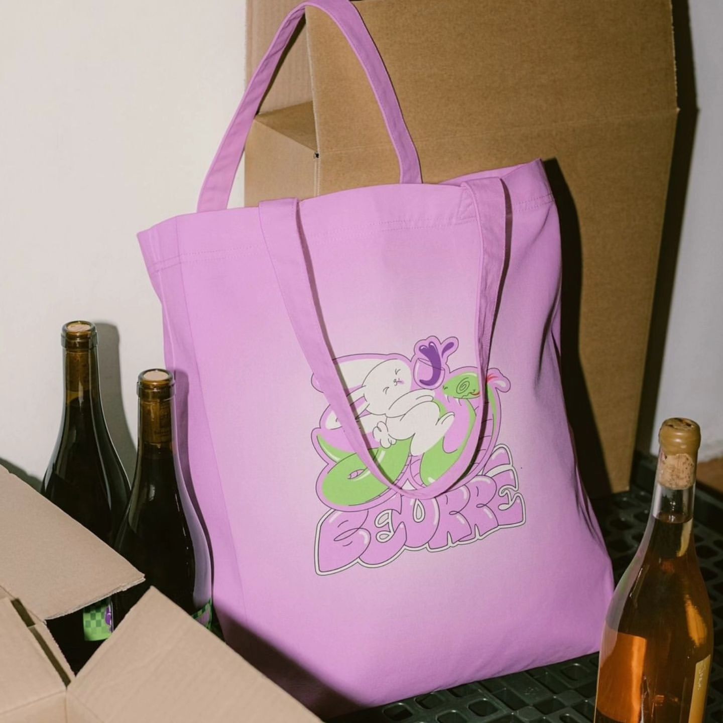 Pink tote bag featuring a cartoon bunny and stylized text, surrounded by wine bottles and cardboard boxes, creating a quirky scene.