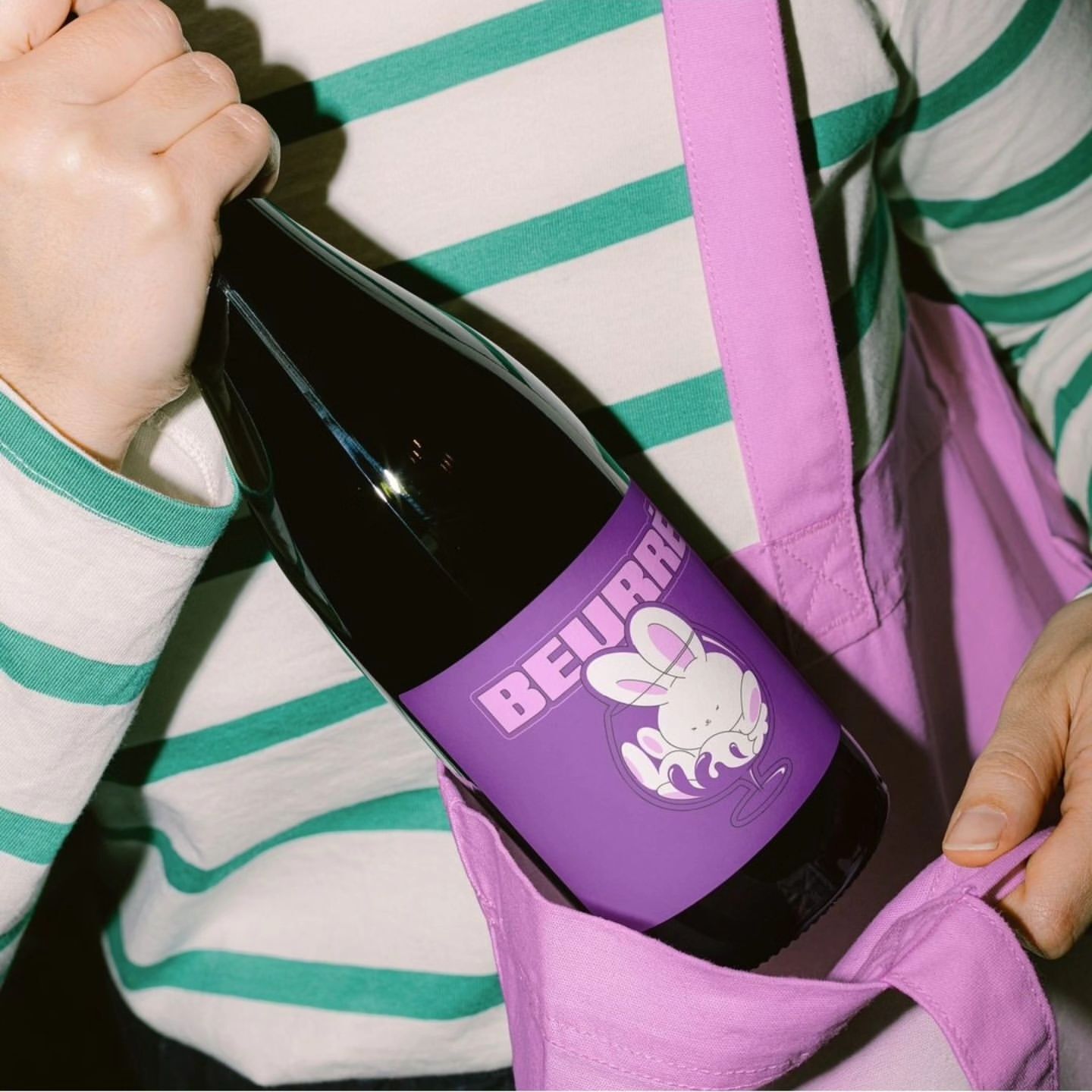 Dark-colored bottle with purple label featuring cartoon rabbit being held in a lilac tote bag. Person in a striped shirt shown.