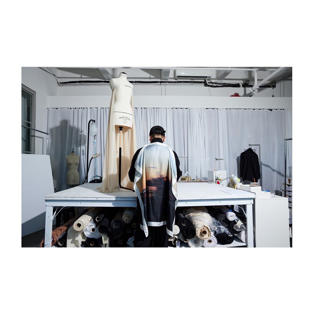 Fashion designer at work in studio with dress form, sewing table with fabric rolls, bright lighting, and neutral color palette.