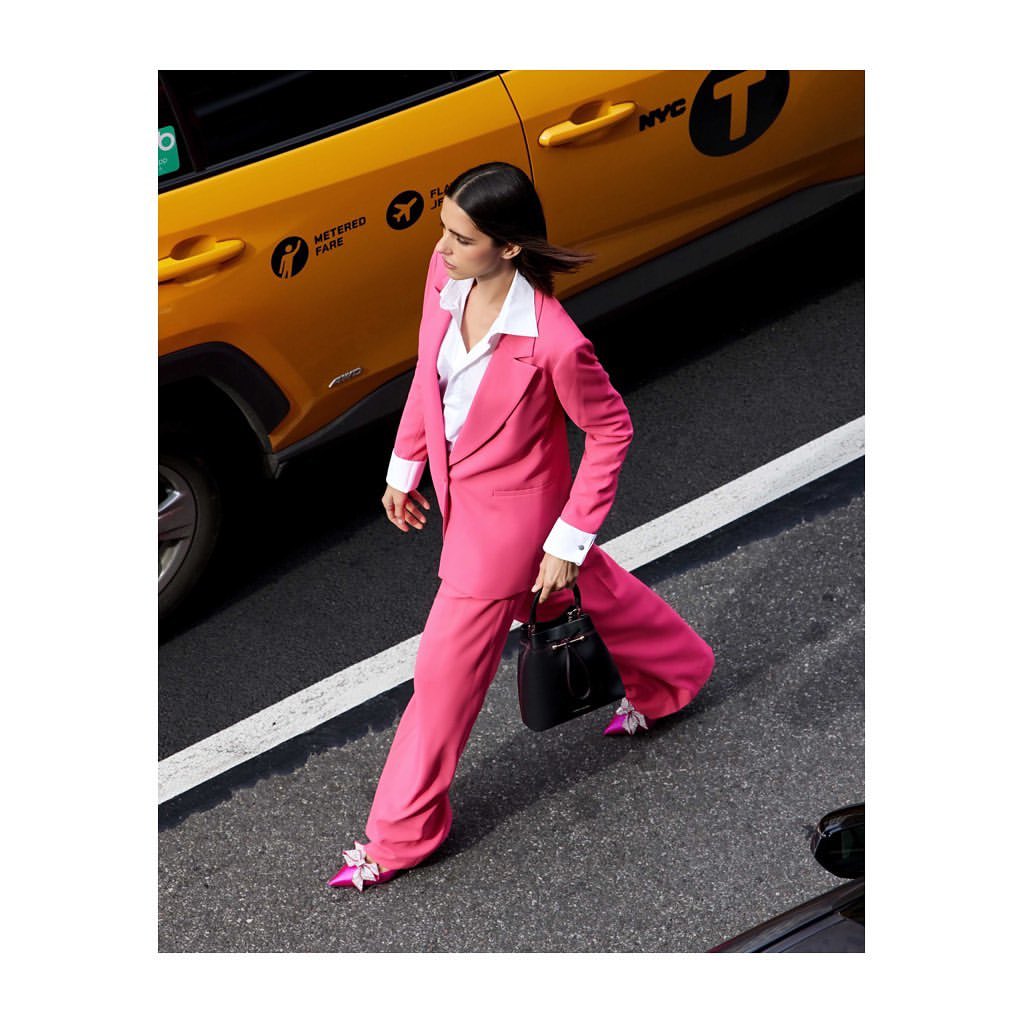 Woman in pink suit walks past yellow taxi in NYC. Stylish professional with modern handbag and matching shoes. Fashion editorial photography.