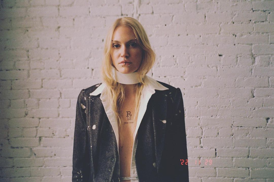 Woman in a blazer with white undershirt poses indoors against a white brick wall, looking at the camera.