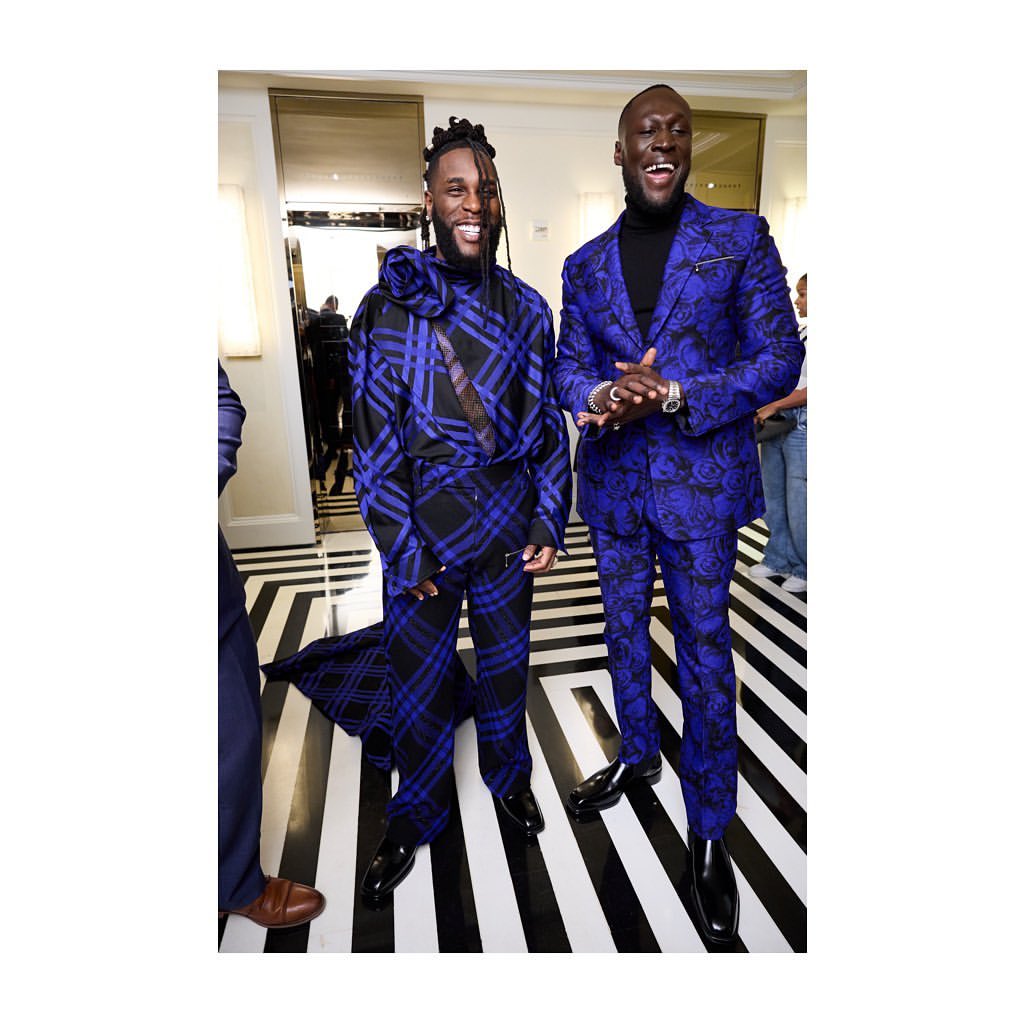 Two men pose in patterned suits. One has a blue plaid pattern and the other has a floral pattern. Black and white striped floor.
