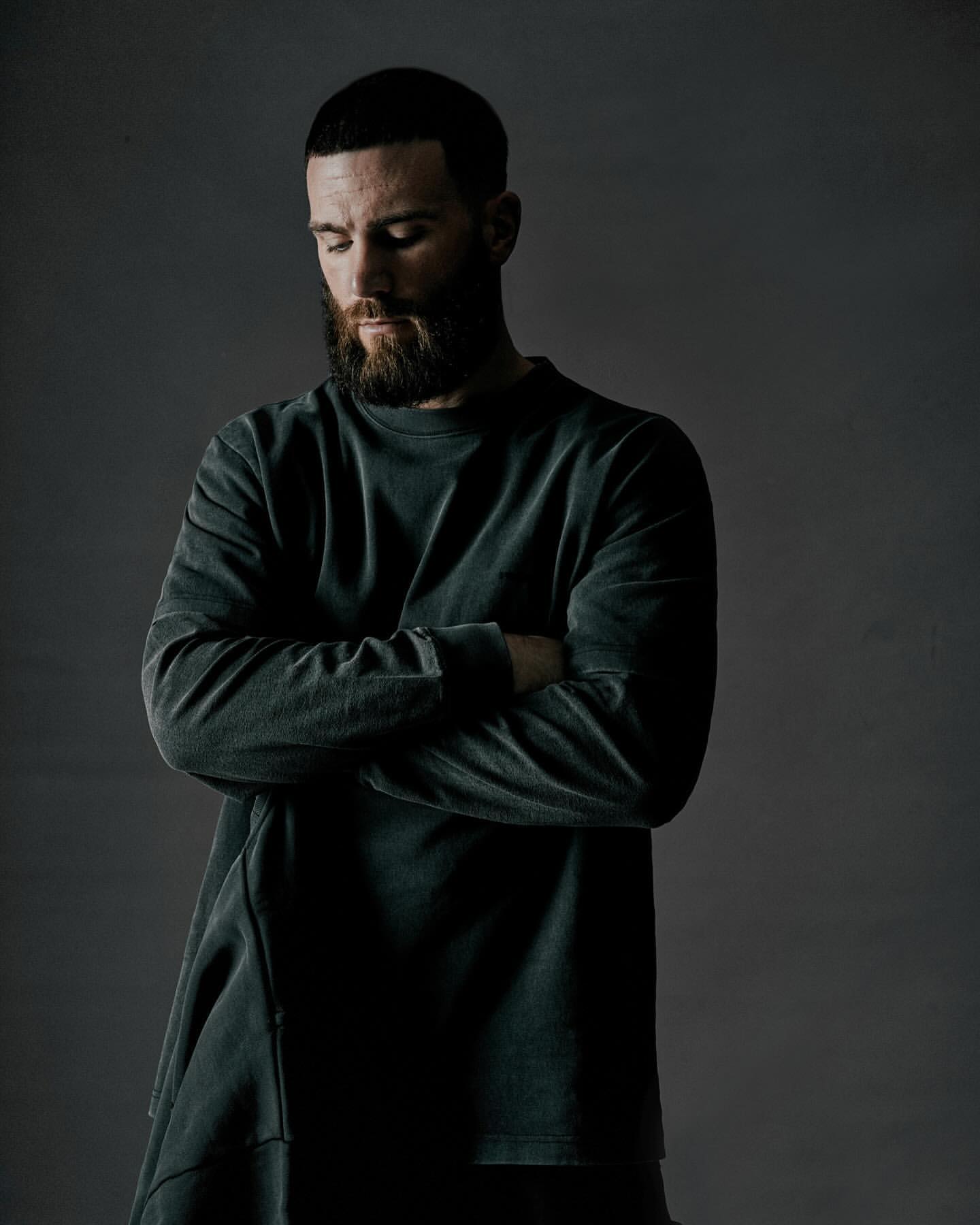 Man with beard wearing a dark long-sleeve sweatshirt with arms crossed against a grey studio backdrop