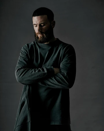 Man with beard wearing a dark long-sleeve sweatshirt with arms crossed against a grey studio backdrop