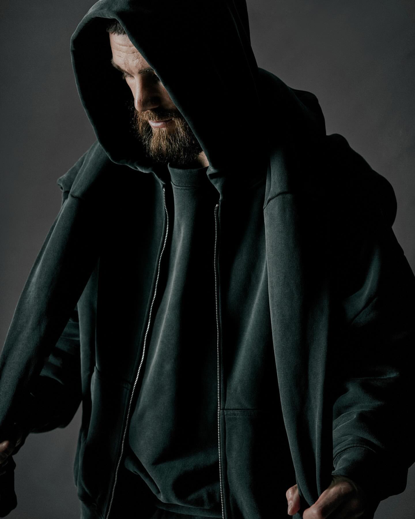 Man wearing a dark hooded sweatshirt with his head bowed down against a neutral grey studio background
