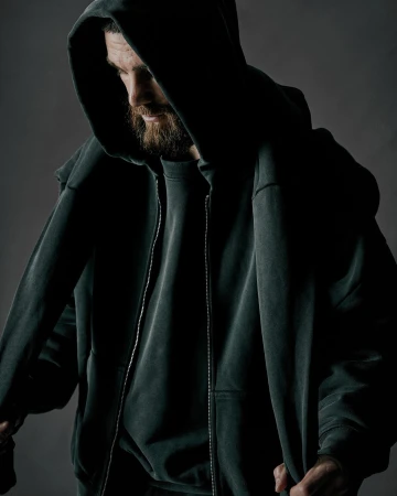 Man wearing a dark hooded sweatshirt with his head bowed down against a neutral grey studio background