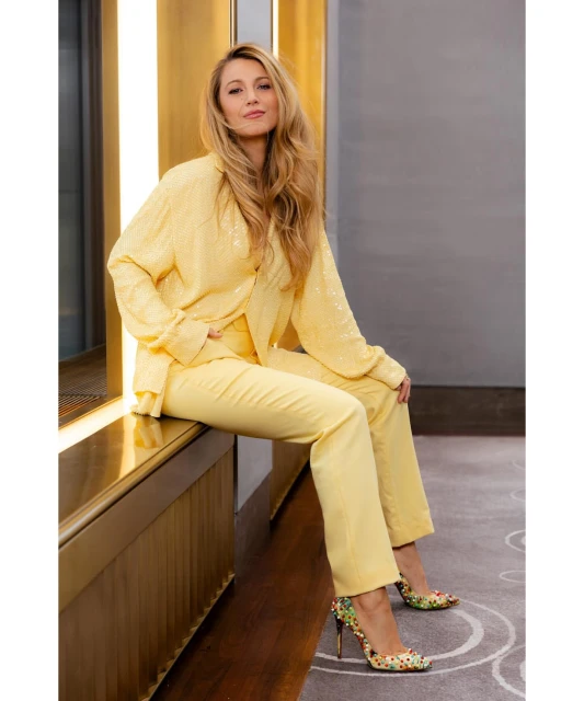 Blonde woman in yellow suit sitting indoors with floral heels. The image captures the stylish attire and elegant pose of the subject.