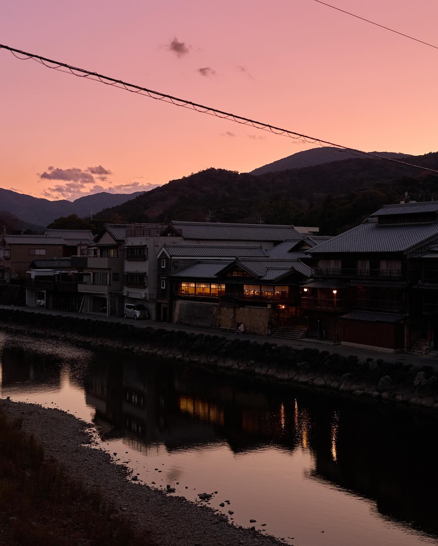Dusk view of a reflective river winding through a Japanese town, silhouetted mountains and a vibrant pink and purple sky in the background.