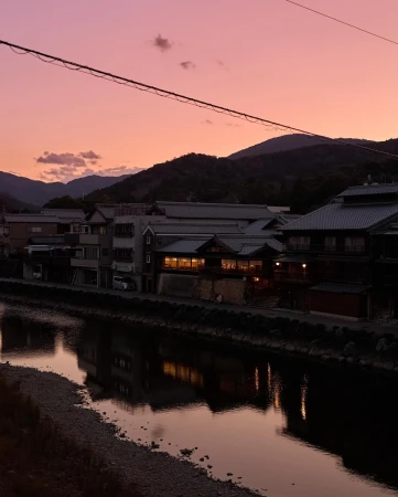 Dusk view of a reflective river winding through a Japanese town, silhouetted mountains and a vibrant pink and purple sky in the background.