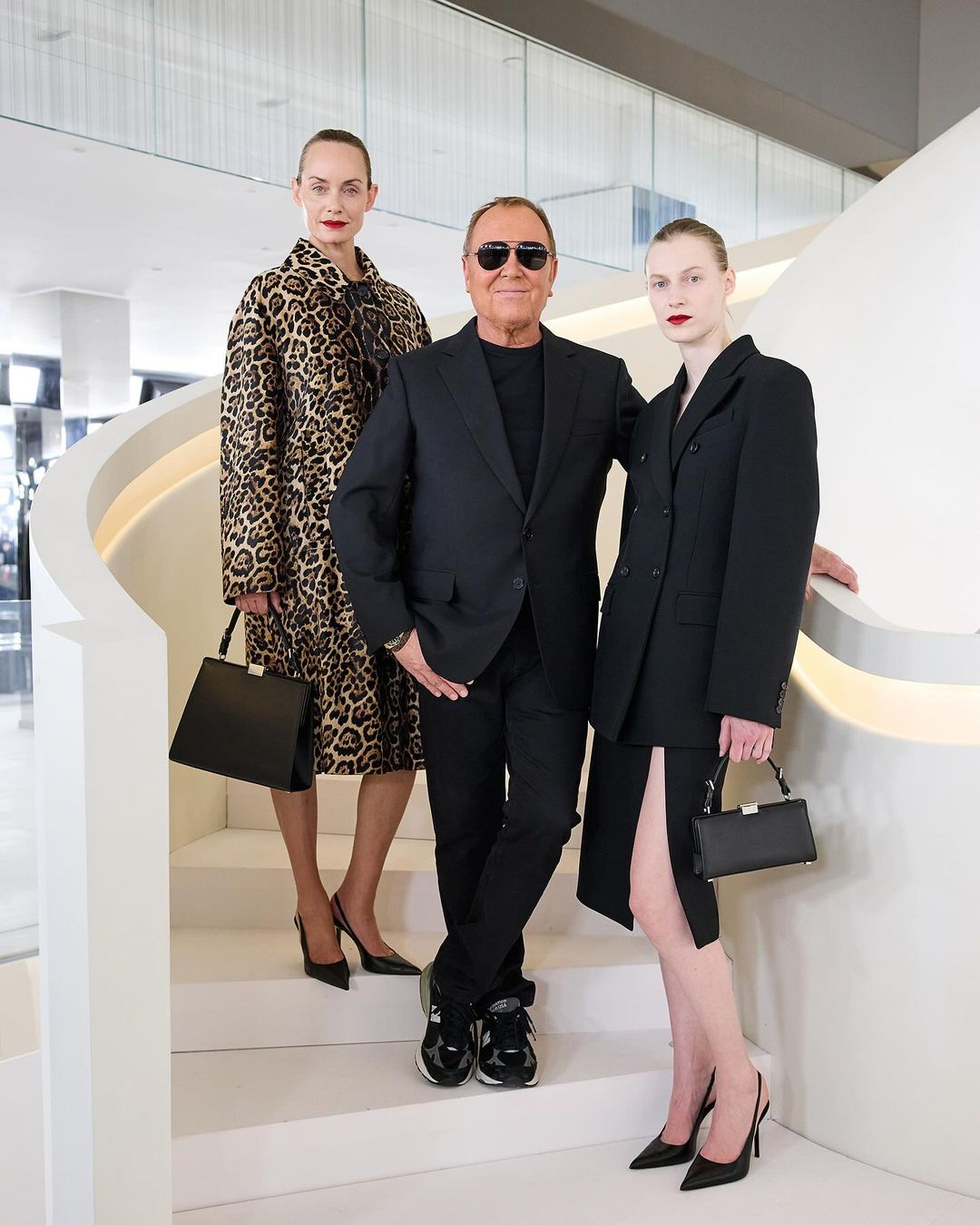 Michael Kors poses between two female fashion models wearing stylish blazers and holding handbags, standing on white staircase.