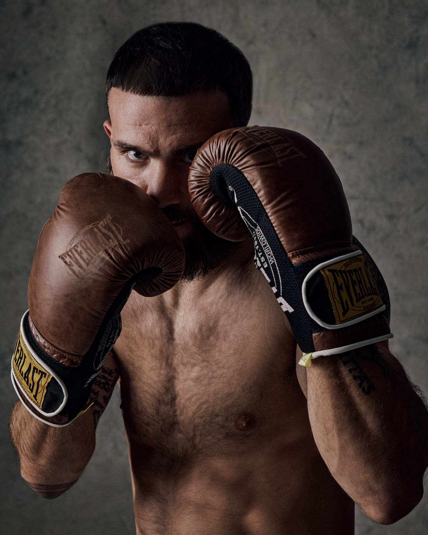 Close-up portrait of a bearded adult male boxer with boxing gloves, posing against a textured grey backdrop in a photo studio setting.