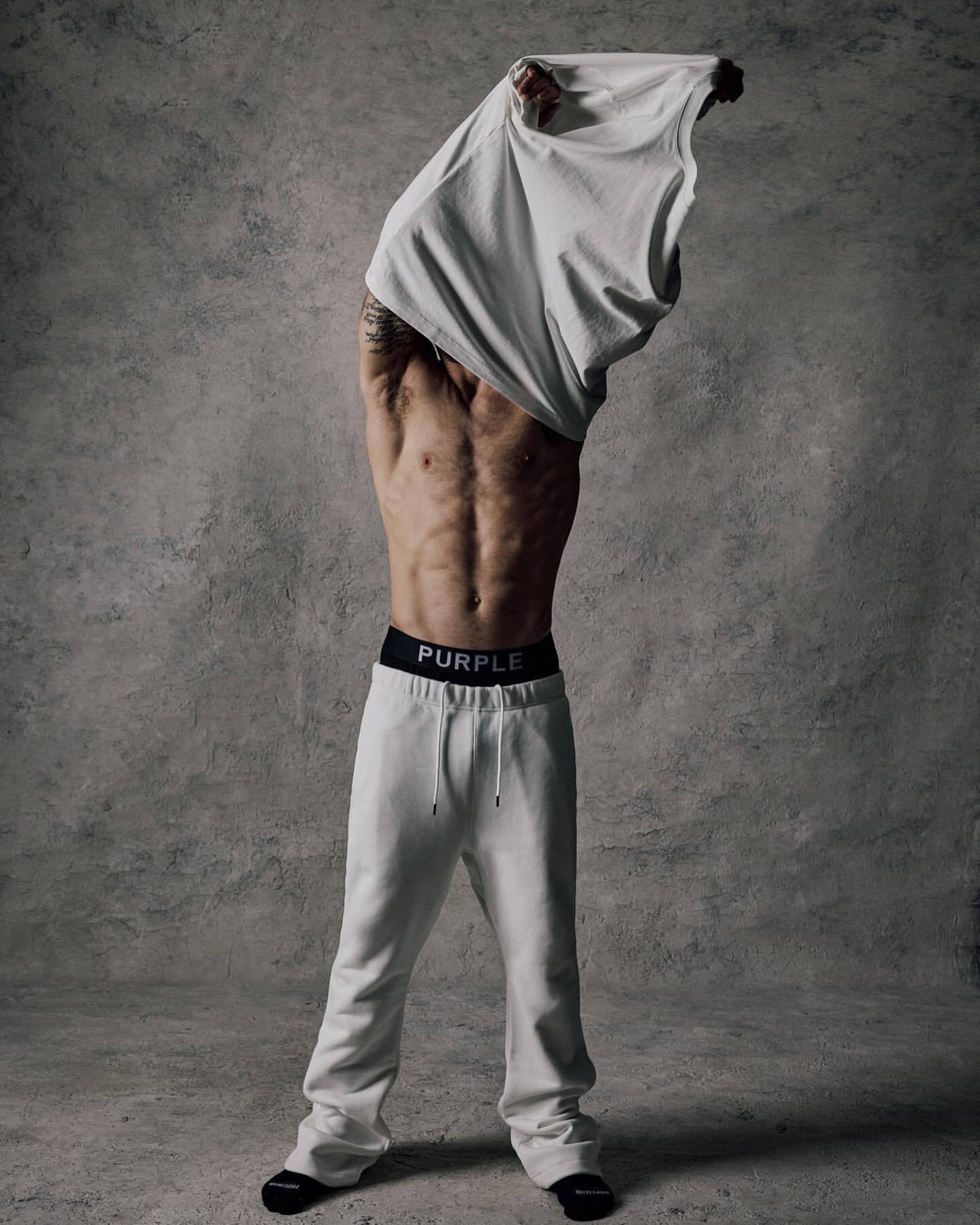 An athletic adult male takes off a white t-shirt, revealing his torso, in an indoor studio with a textured grey background and natural lighting.