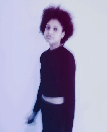 Portrait of a young woman with a dark afro hairstyle wearing a long sleeve black top against a light background