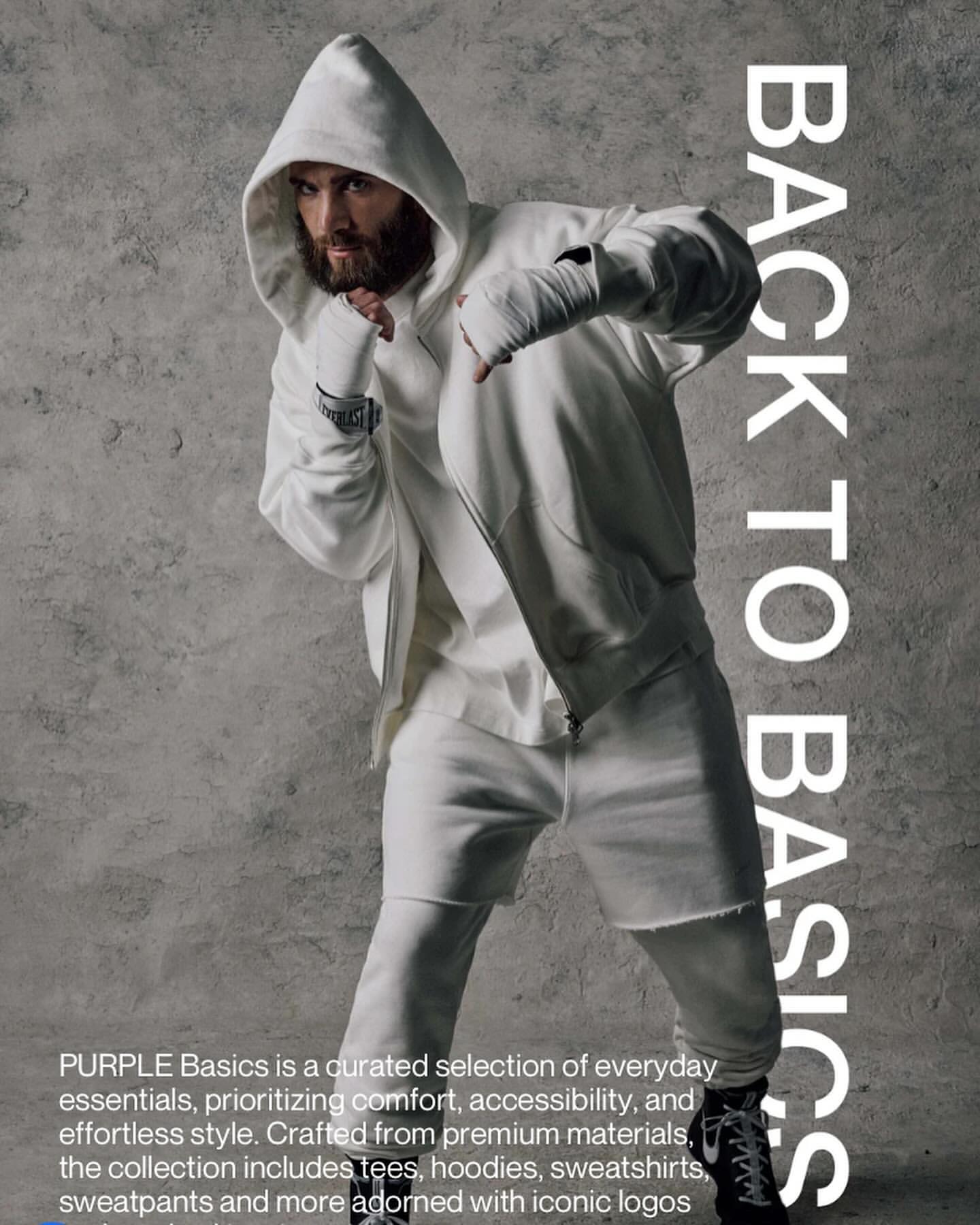 Man in white hoodie and sweatpants with hand wraps striking a boxer pose against a mottled grey background, looking at the camera.