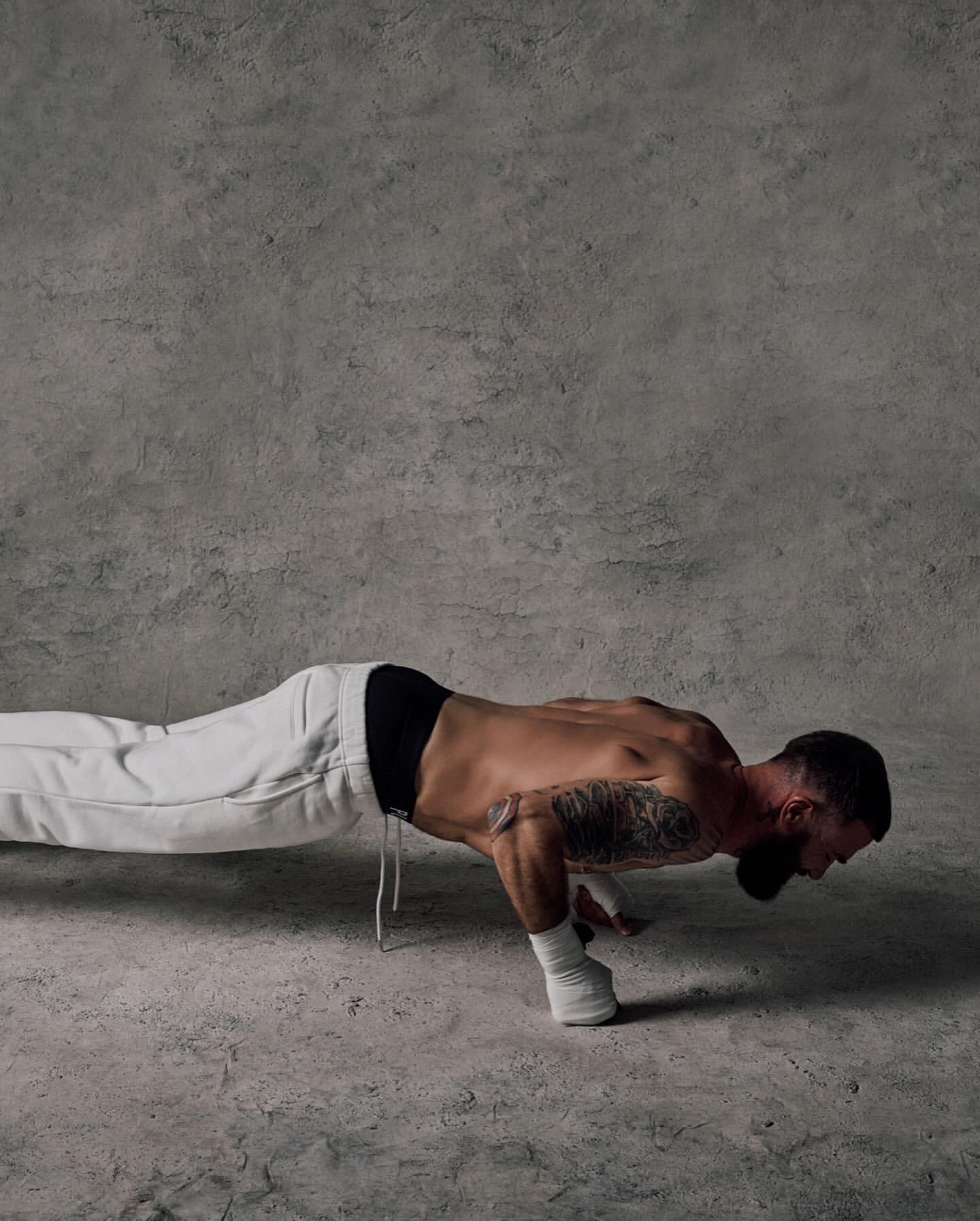 Man doing a one-handed push-up in a studio. He wears white sweatpants and has a tattoo on his arm. Grey concrete background and floor.
