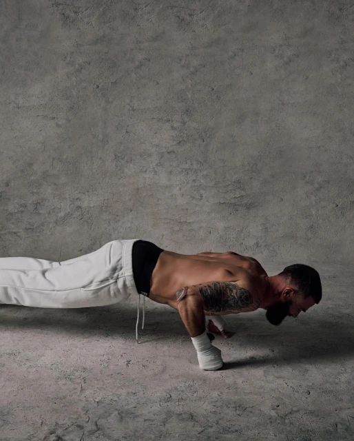 Man doing a one-handed push-up in a studio. He wears white sweatpants and has a tattoo on his arm. Grey concrete background and floor.